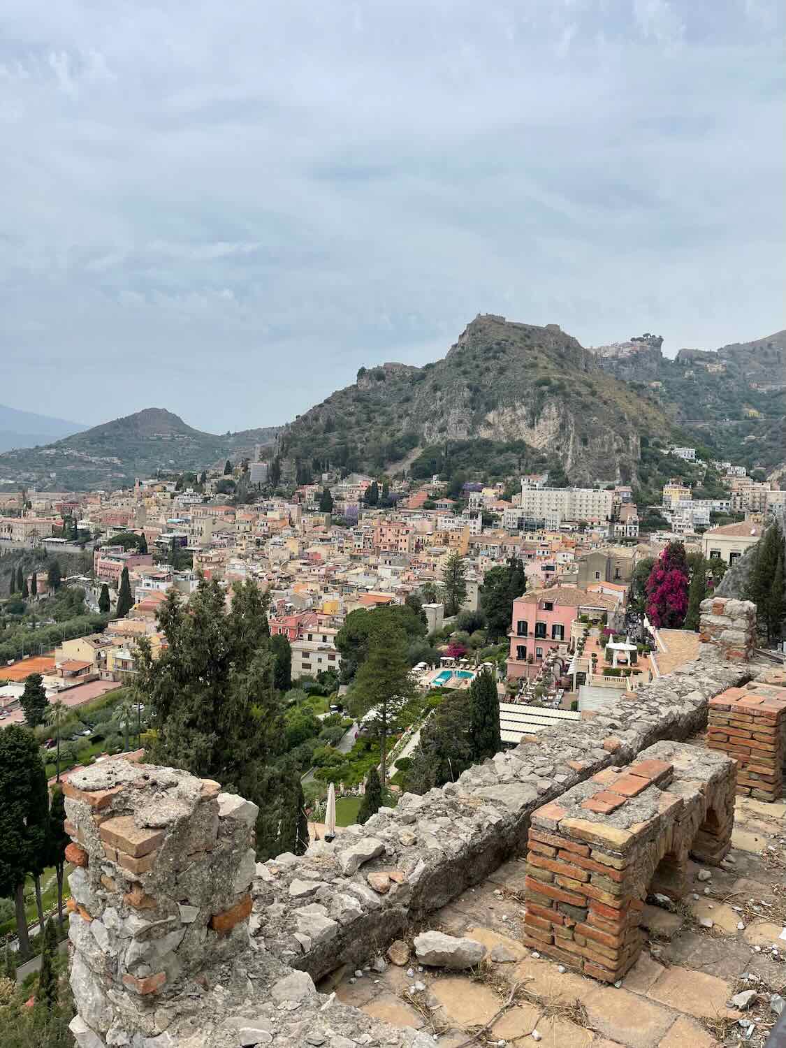 A scenic view of Taormina, Sicily, showcasing a mix of historic ruins in the foreground and the sprawling town nestled among green hills and mountains in the background. The buildings are predominantly low-rise, with a blend of colorful and neutral tones, giving a picturesque and charming appearance.