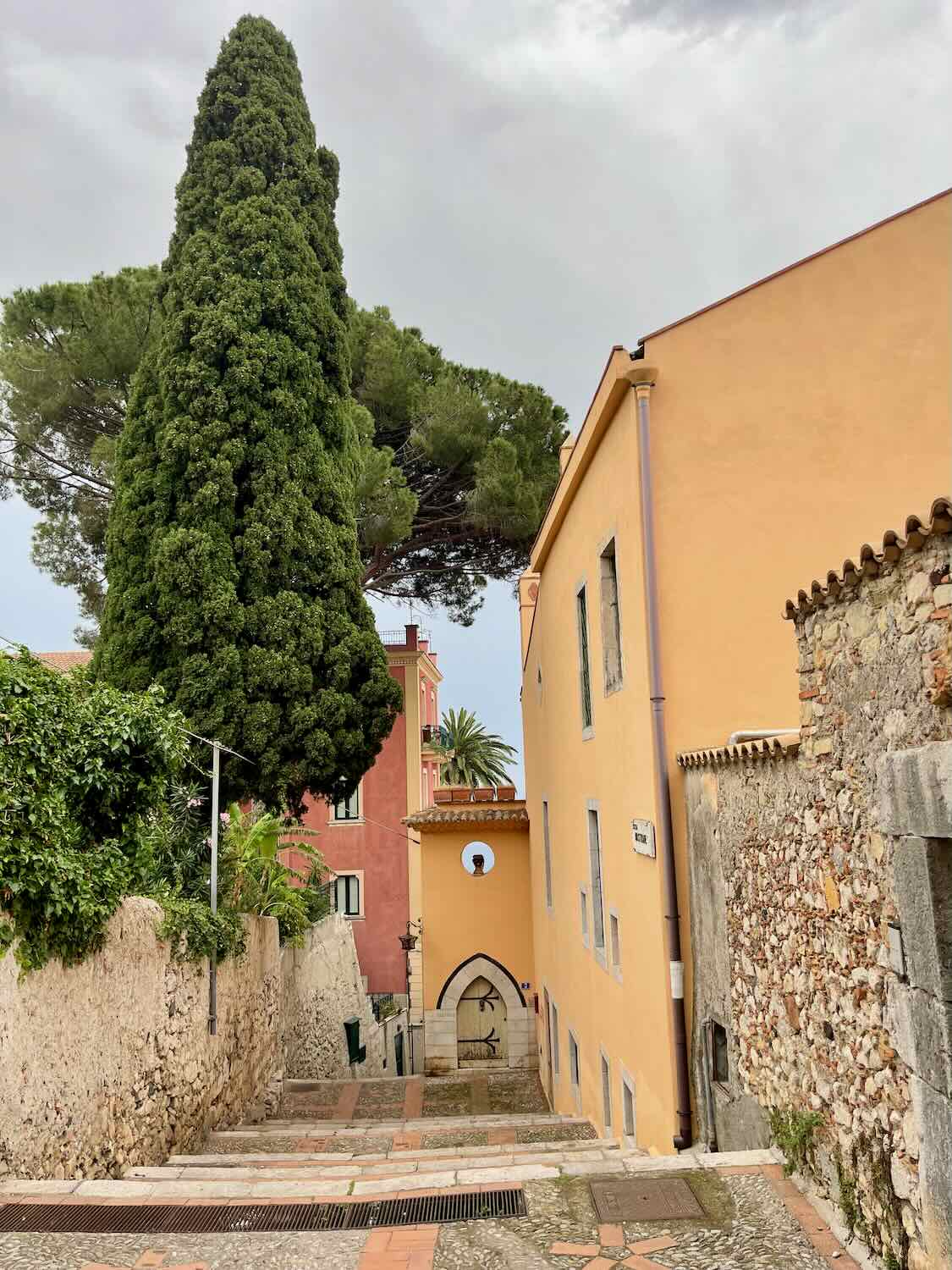 A picturesque view of a church and surrounding buildings in Taormina, framed by a tall cypress tree and stone walls.