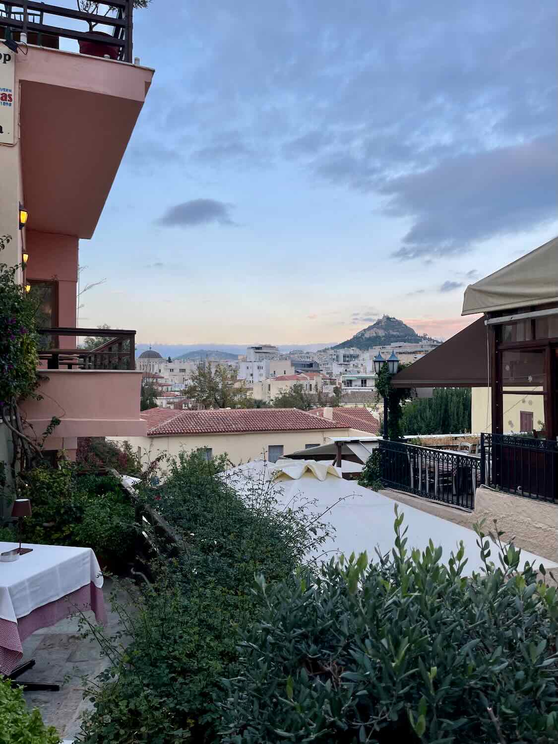 A view of Athens from a balcony at dusk, with Mount Lycabettus visible in the distance. The foreground features a table with a checkered tablecloth and lush greenery.