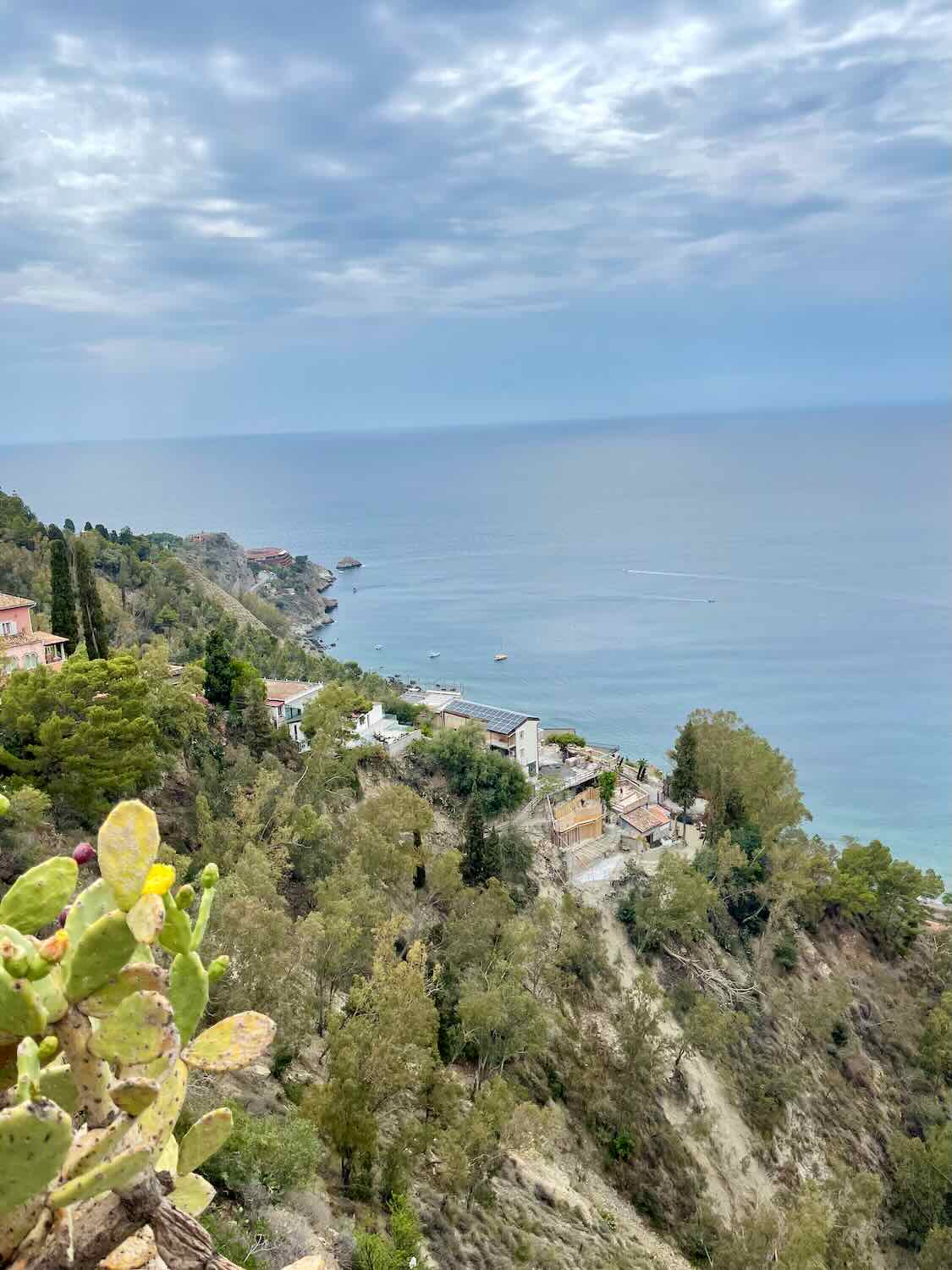 A coastal view from Taormina, Sicily, overlooking the Mediterranean Sea. The image captures the rugged coastline, with green vegetation and a few scattered buildings along the shore. The sky is partly cloudy, creating a serene and calm atmosphere.