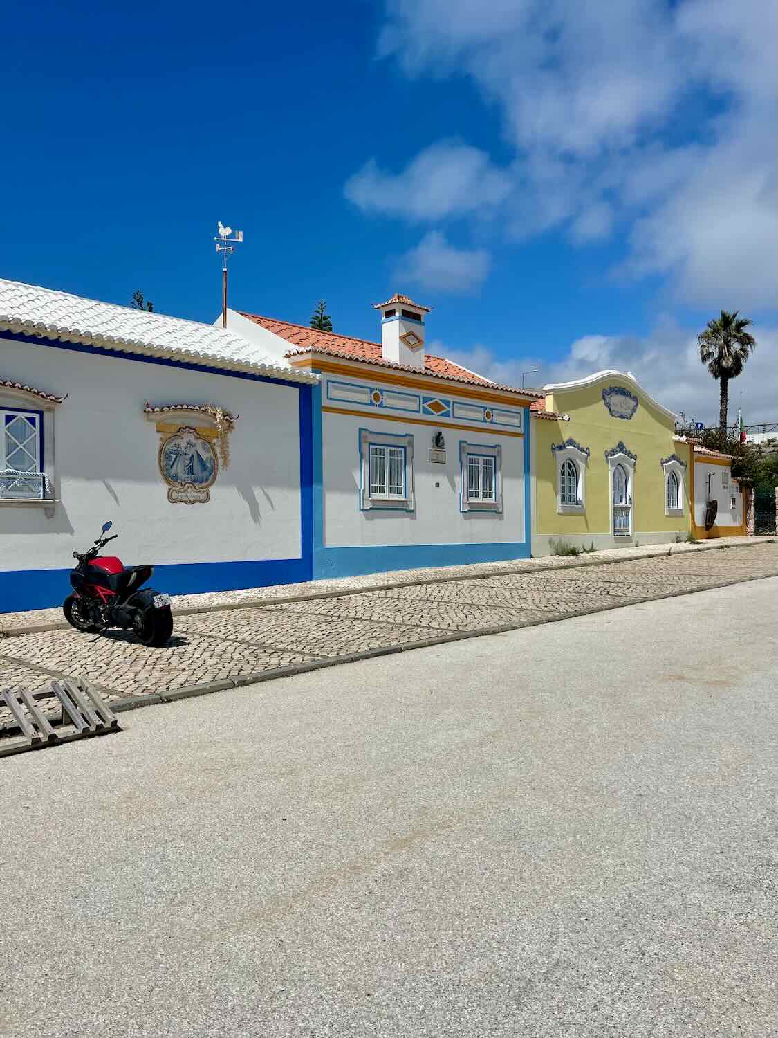 A bright and sunny street scene in Ericeira featuring colorful traditional Portuguese cottages. The buildings have white walls with blue and yellow trim, red-tiled roofs, and decorative accents. A red motorcycle is parked on the cobblestone street under a clear blue sky.