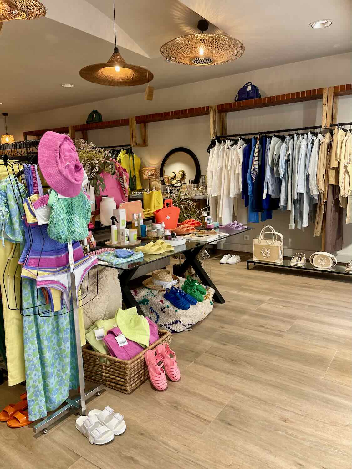 The interior of a trendy boutique featuring vibrant and colorful clothing, accessories, and other lifestyle products. The store has a cozy and contemporary design with wooden flooring and stylish lighting fixtures. Various items, including swimsuits, hats, and skincare products, are neatly arranged on racks and tables, creating an inviting shopping experience.