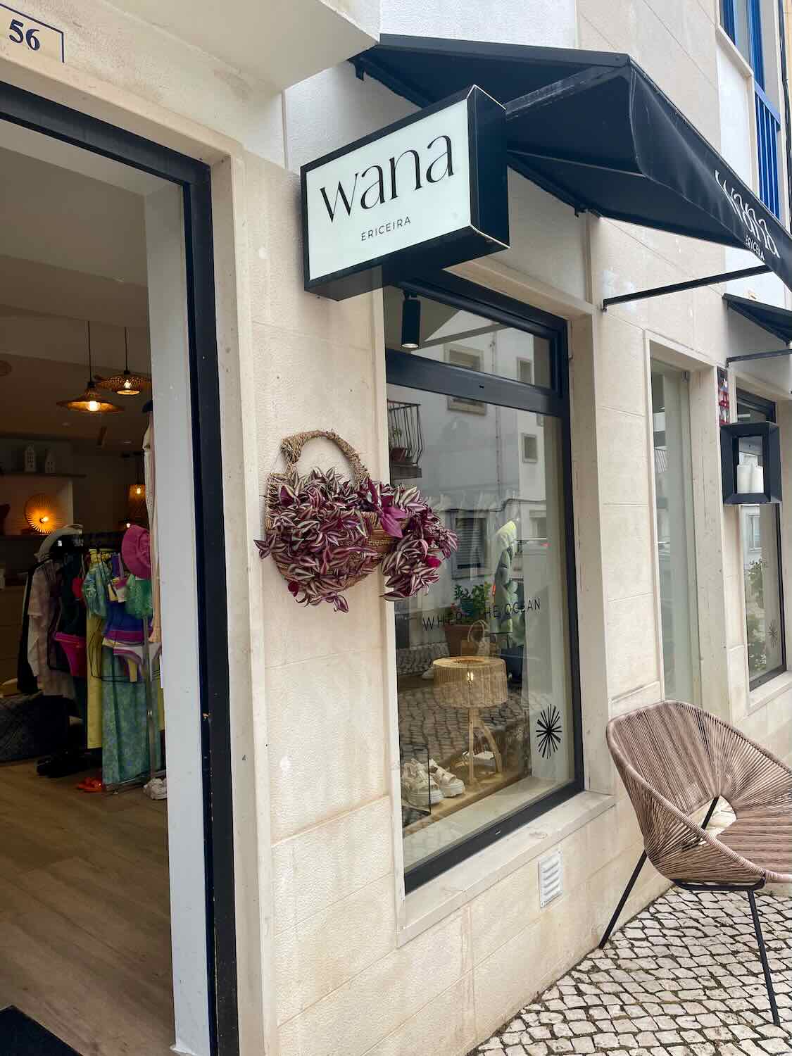 The exterior of a boutique named "Wana" located in Ericeira. The store's sign is prominently displayed above the entrance, and a woven basket with decorative items hangs near the door. Through the windows, the interior displays a range of fashionable clothing and accessories. The entrance area is paved with cobblestones, contributing to the quaint and charming atmosphere.