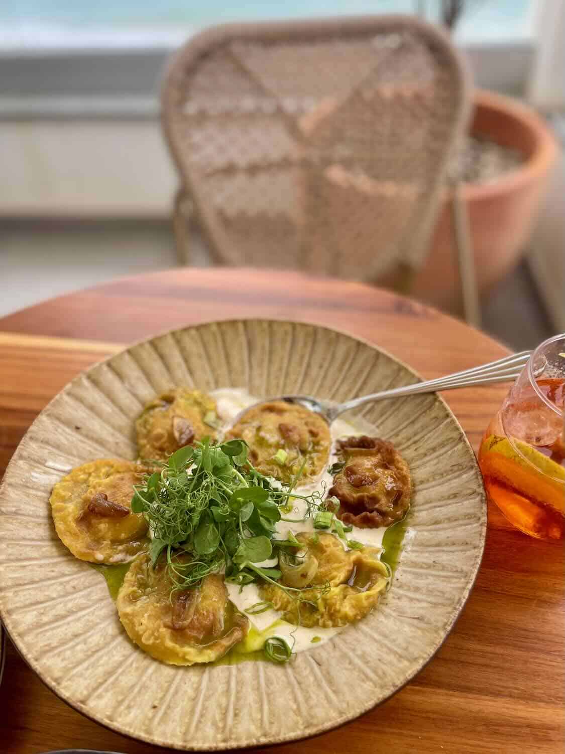 A plate of beautifully presented ravioli, garnished with fresh greens and accompanied by a beverage in a glass with slices of orange. The dish is set on a wooden table with a woven chair in the background.