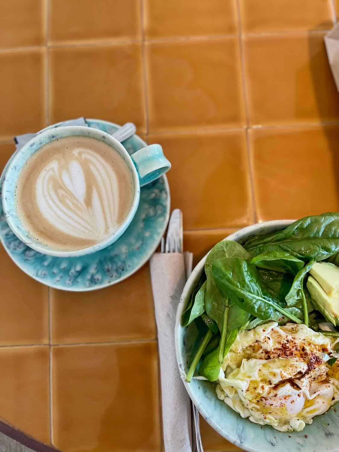 A cozy breakfast setting with a cup of latte featuring a heart-shaped foam art and a bowl of fresh salad with greens, avocado, and poached eggs. The items are placed on a tiled table with a set of cutlery wrapped in a napkin.