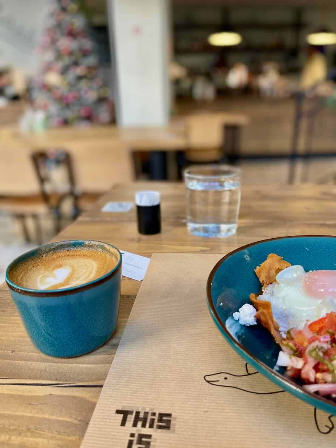 A table with a teal cup of cappuccino, a glass of water, and part of a breakfast dish with poached eggs, salsa, and yogurt. The background features a cozy café interior with a Christmas tree visible in the distance.