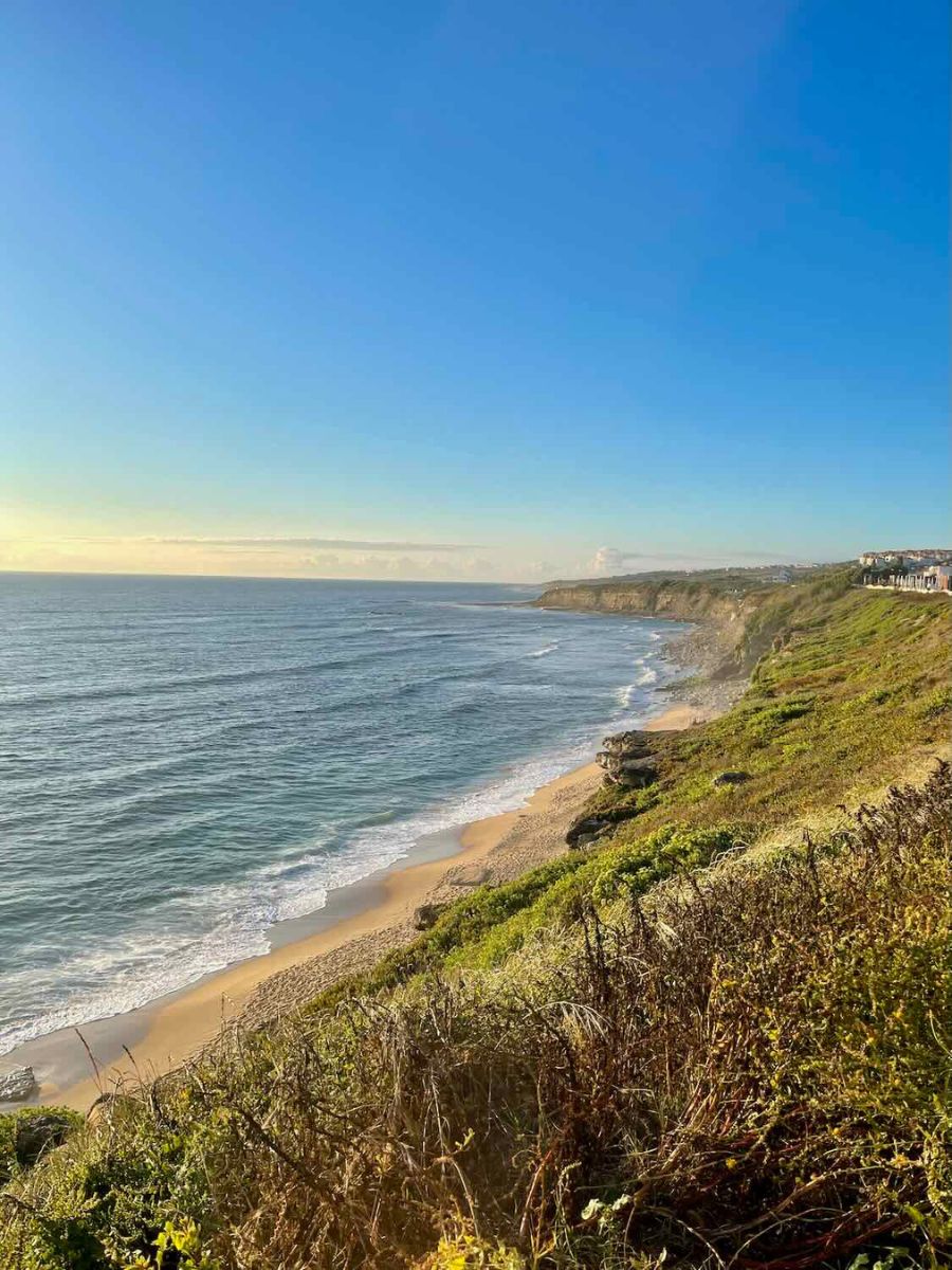 View from a cliff overlooking a sandy beach in Ericeira, Portugal, with the ocean waves gently lapping against the shore. The sun is shining brightly in a clear blue sky, casting a warm light over the scene. The coastline stretches into the distance, bordered by green vegetation and rocky formations.