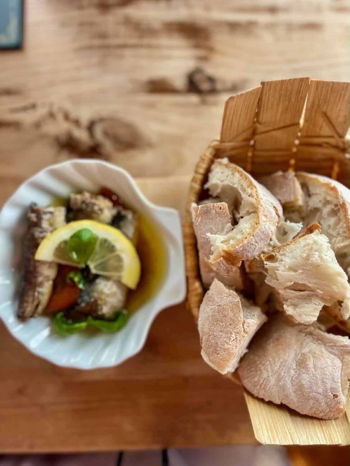 A basket of crusty bread served alongside a dish of marinated sardines garnished with a lemon slice and basil, set on a wooden table at Mar das Latas Wine & Food.