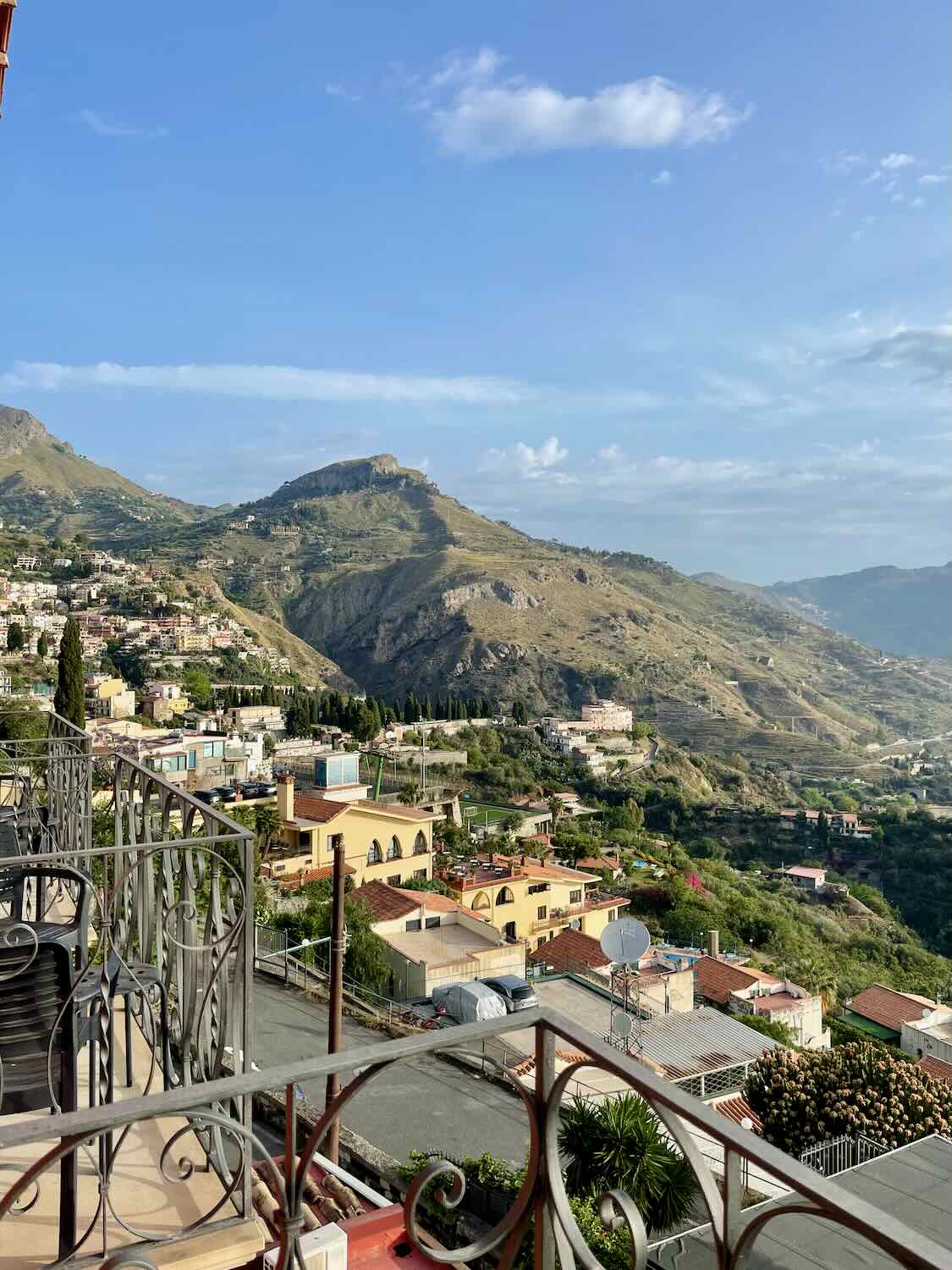 Scenic view from a balcony in Taormina overlooking the town and surrounding mountains under a clear blue sky.