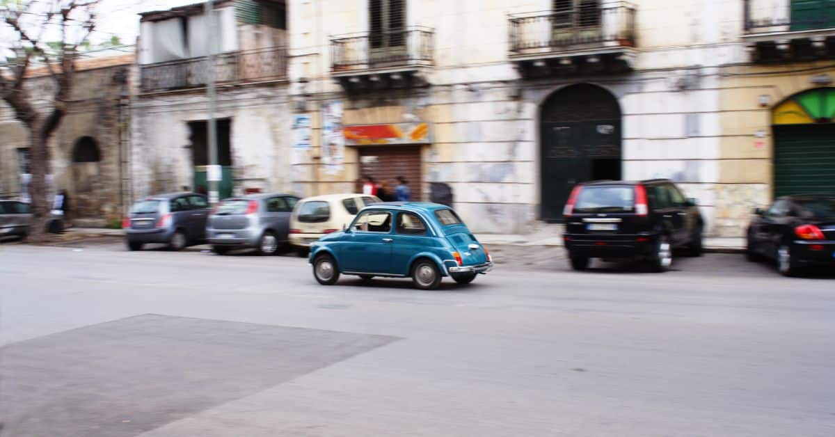 A blurred photograph of a small blue vintage car driving down a street in Sicily, passing by other parked cars and historic buildings with faded paint. The scene captures the essence of everyday life in a Sicilian town, with a sense of movement and charm from the classic car.