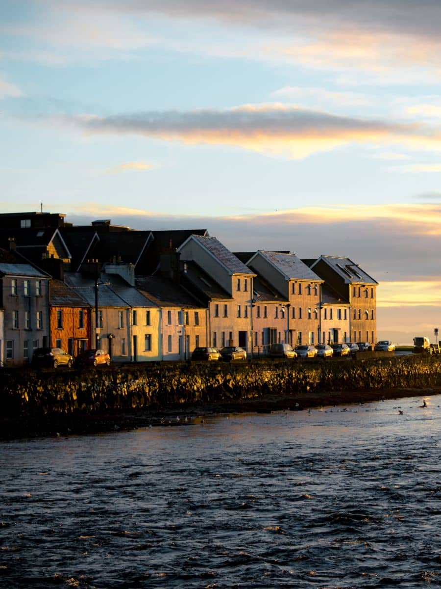 Row of pastel-colored houses along a waterfront in Galway, Ireland, with the sun setting in the background, casting a warm golden light on the buildings.