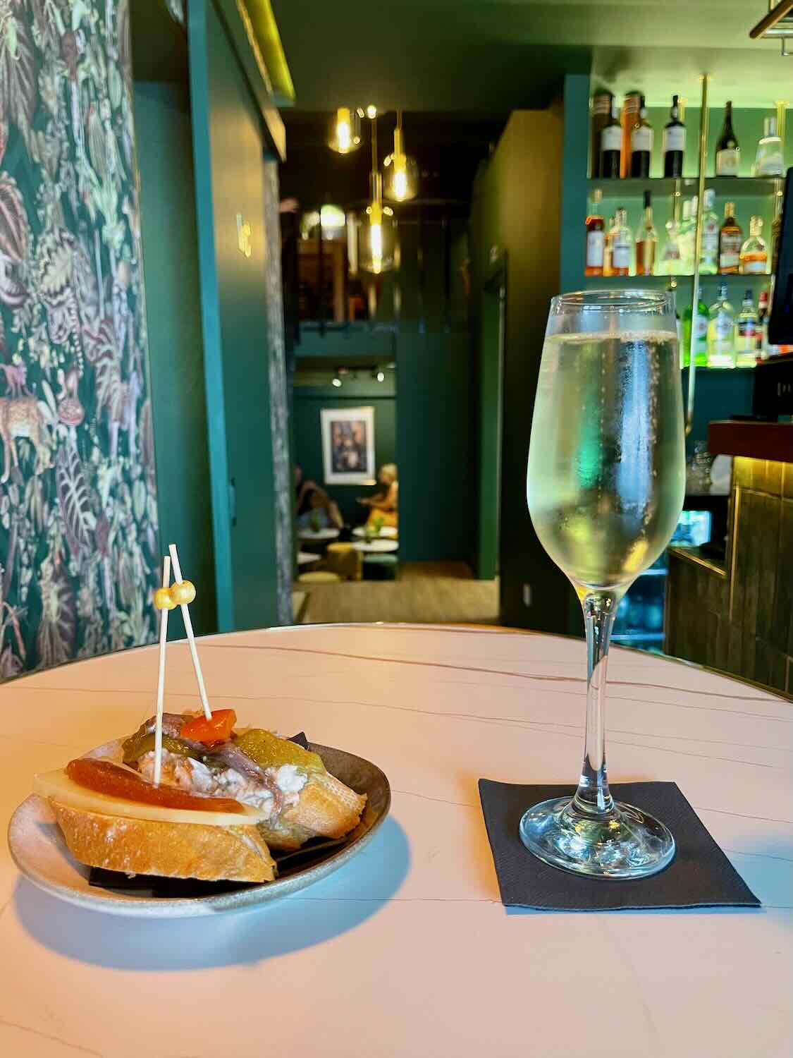 A bar scene with a sparkling wine glass and a small plate of tapas on a round table. The background features a stylish bar with various bottles and a dimly lit, cozy atmosphere.