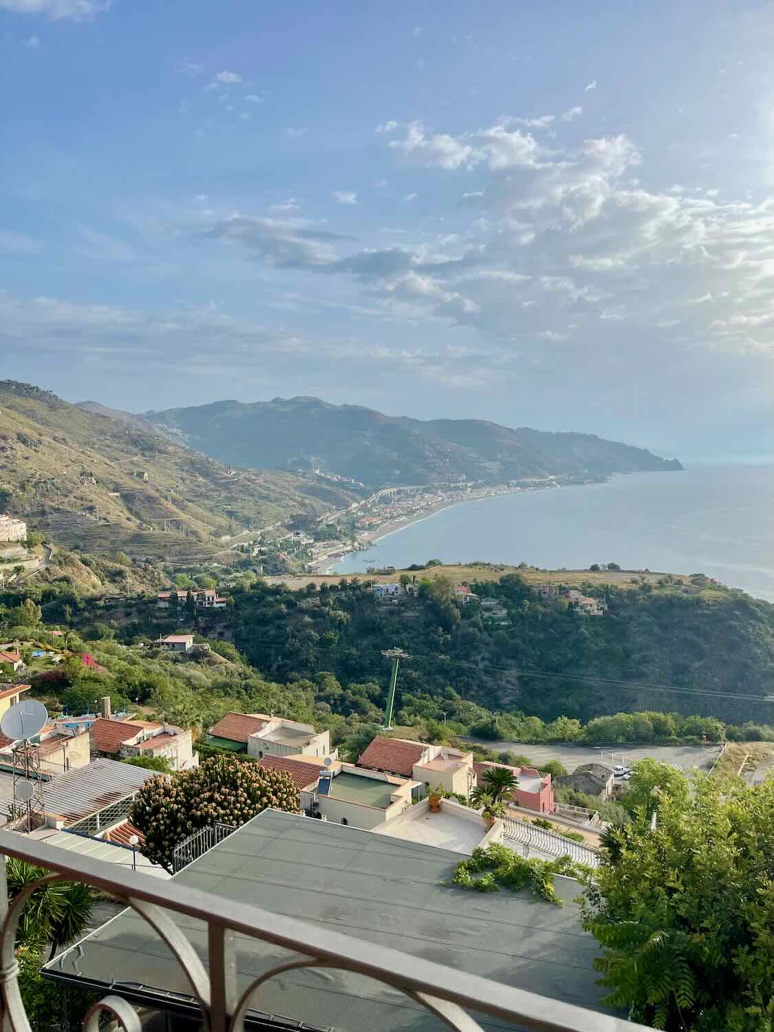 A stunning view from a balcony in Taormina overlooking the town, the coastline, and surrounding mountains under a clear blue sky.