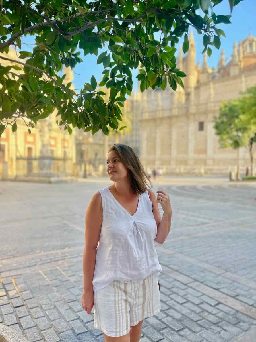 The photo shows a woman standing outdoors in Seville, with a peaceful and contemplative expression. She is dressed in a casual, light-colored outfit, perfect for a warm day. The background includes a tree branch with green leaves framing the top of the image, and the architectural details of Seville can be seen in the distance. The scene captures a serene moment, possibly reflecting the calm and beauty of the city.