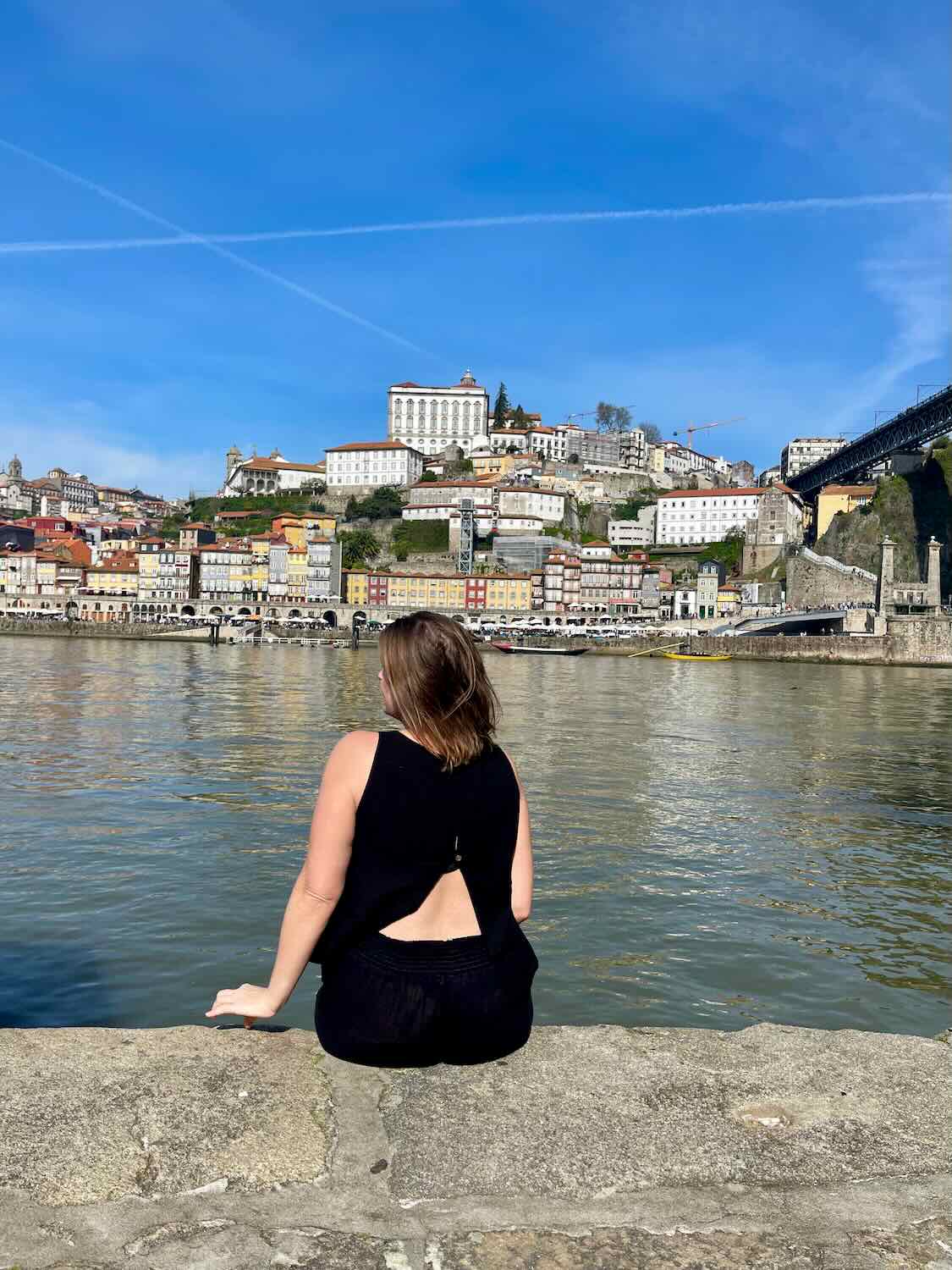 A person in a black outfit sits on the edge of a riverbank in Porto, facing the city's colorful hillside buildings and the Douro River.
