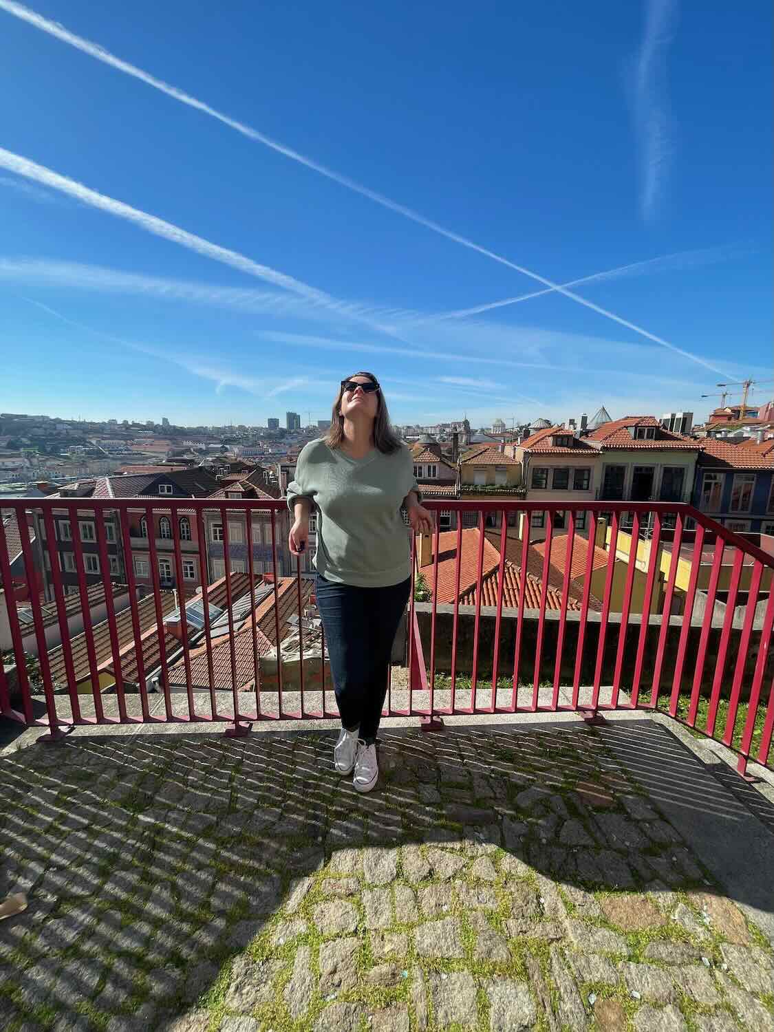 A person stands on a cobblestone terrace in Porto, looking up at the clear blue sky with crisscrossing contrails, surrounded by red-roofed buildings.