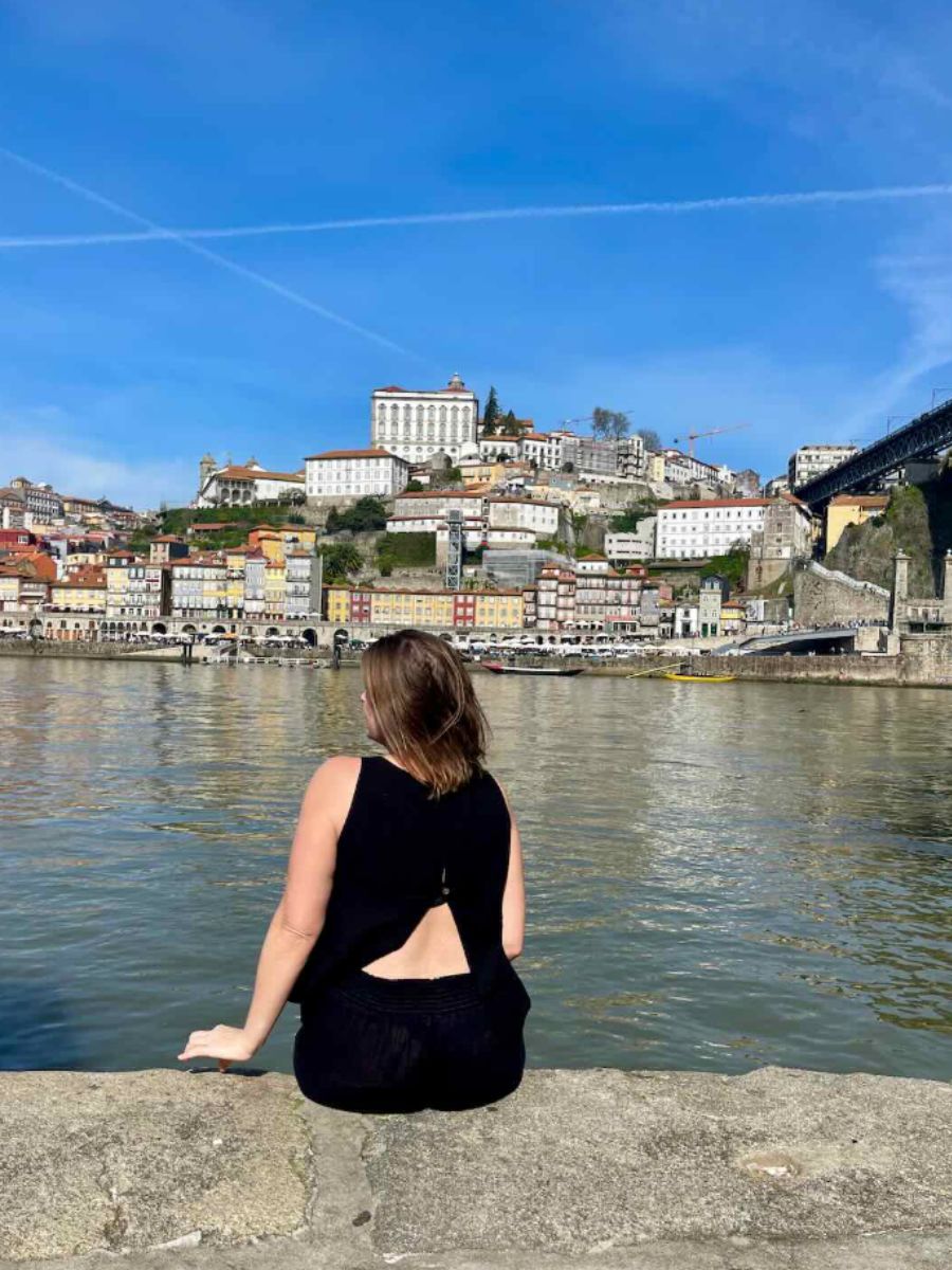 A woman in a black outfit sits on the edge of a riverbank in Porto, facing the city's colorful hillside buildings and the Douro River.