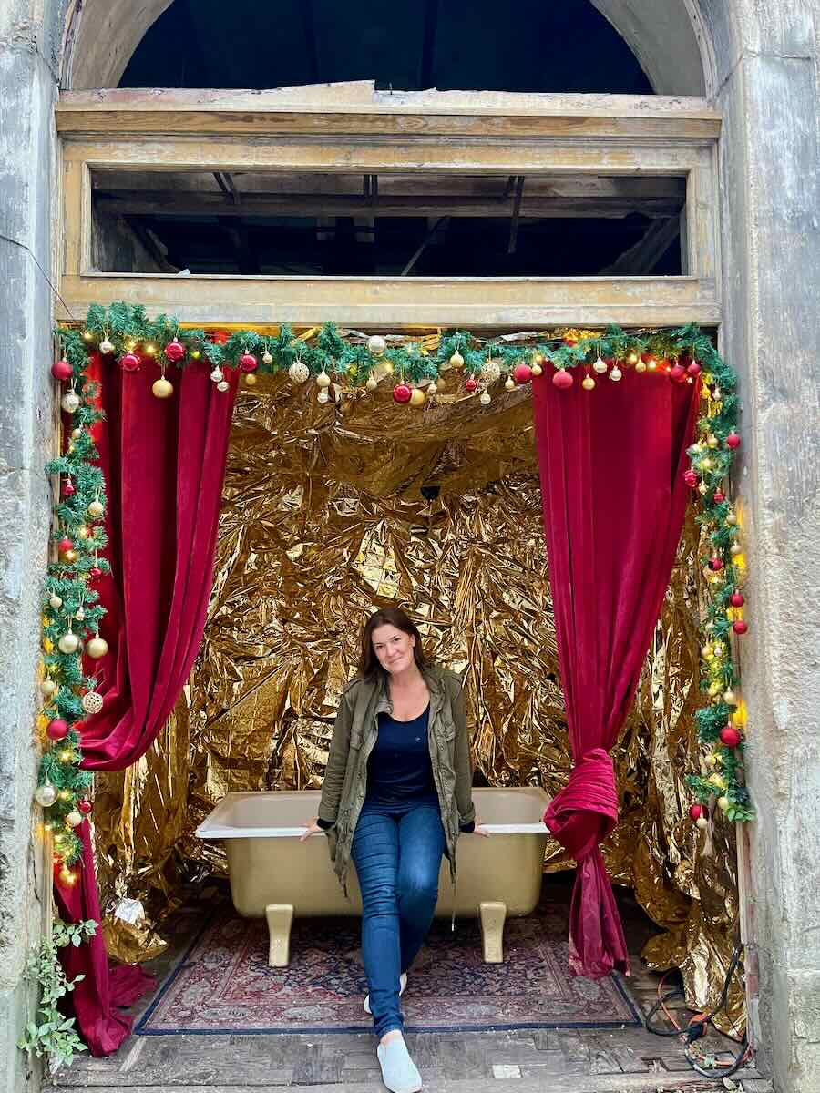 A woman sits on the edge of a vintage bathtub, which is set up in a festive, makeshift photo booth decorated with red curtains, Christmas ornaments, and gold foil in Lisbon, Portugal.