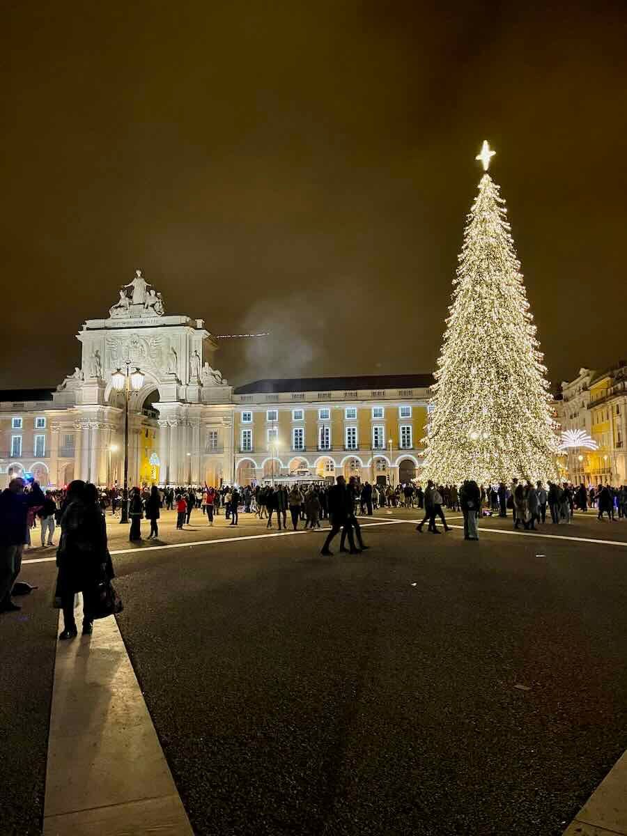 Nighttime view of Praça do Comércio in Lisbon, Portugal, during the Christmas season. The square is illuminated by a towering Christmas tree adorned with white lights and a glowing star on top.