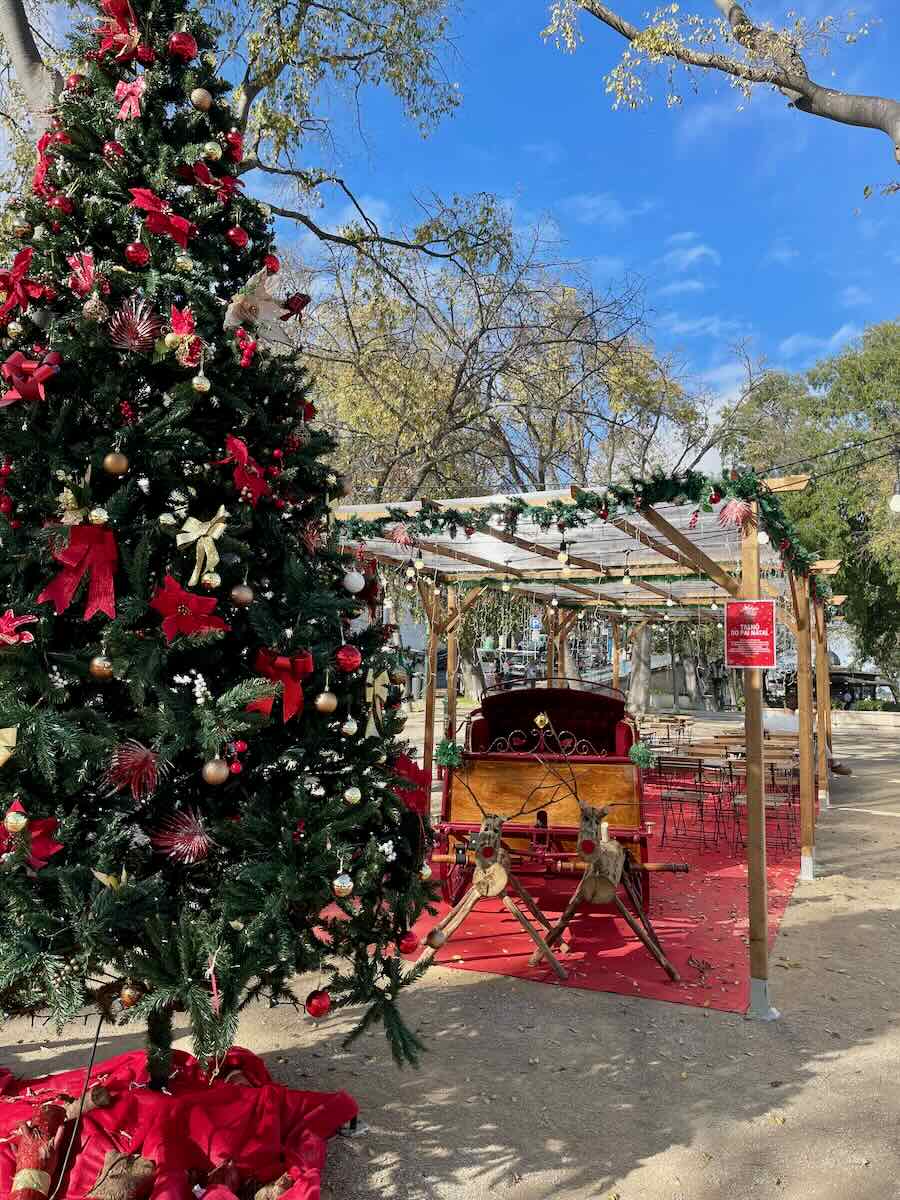 Large Christmas tree decorated with red ribbons and ornaments in a Lisbon Christmas pop-up market, with festive tables and a reindeer sleigh nearby.