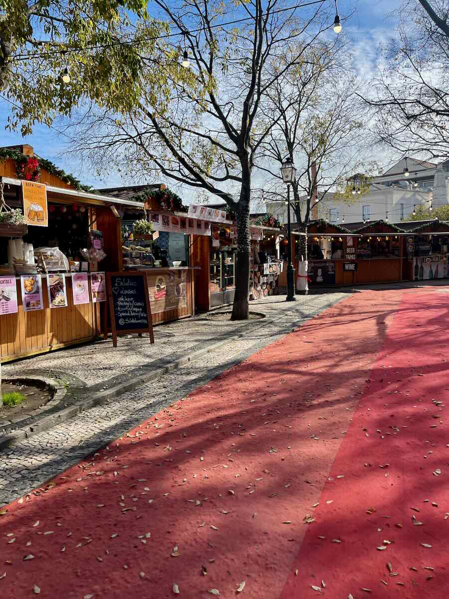 Wooden stalls at a Lisbon Christmas pop-up market along a red pathway, decorated with festive garlands and menu boards showcasing food and drink options.