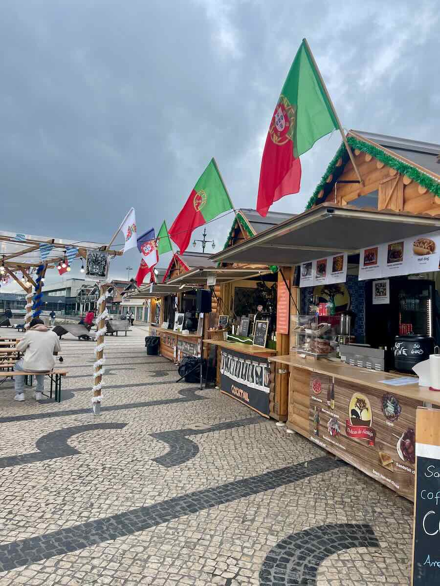 Market stalls decorated with Portuguese flags along the riverfront in Lisbon, with a few visitors seated at tables under a cloudy sky.