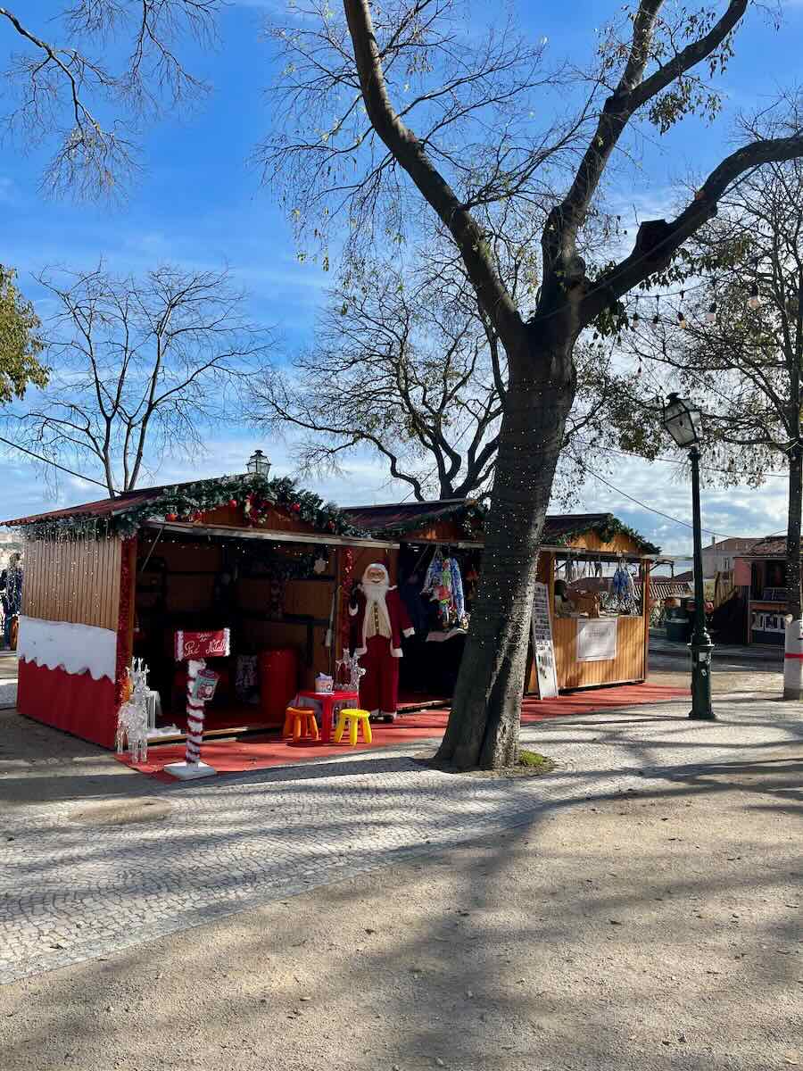 A wooden Christmas market stall in Lisbon decorated with garlands and festive lights, featuring a Santa Claus figure and small stools on a bright winter day.