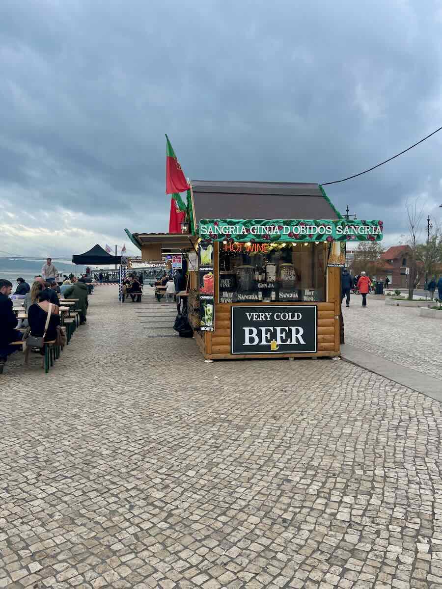 Small wooden booth at a Christmas market in Lisbon offering sangria, hot wine, and beer, with outdoor seating and cloudy skies by the riverfront.