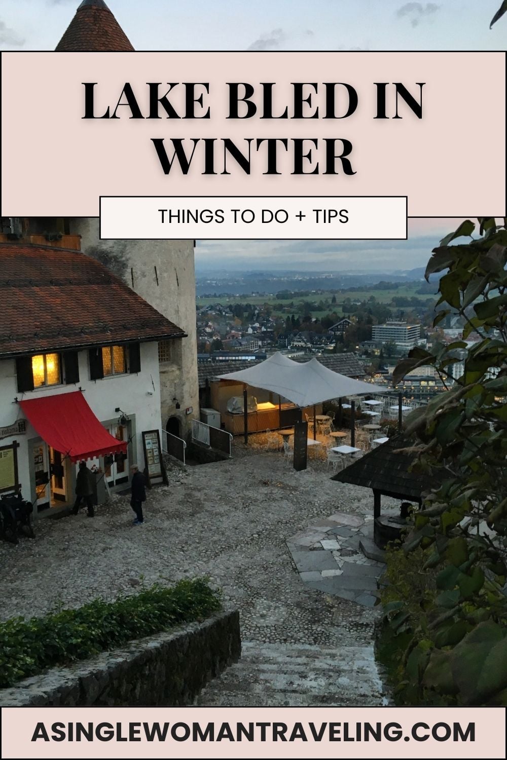 Winter view from Bled Castle overlooking Lake Bled at dusk, with cobblestone courtyard, lit café terrace, and alpine town below.