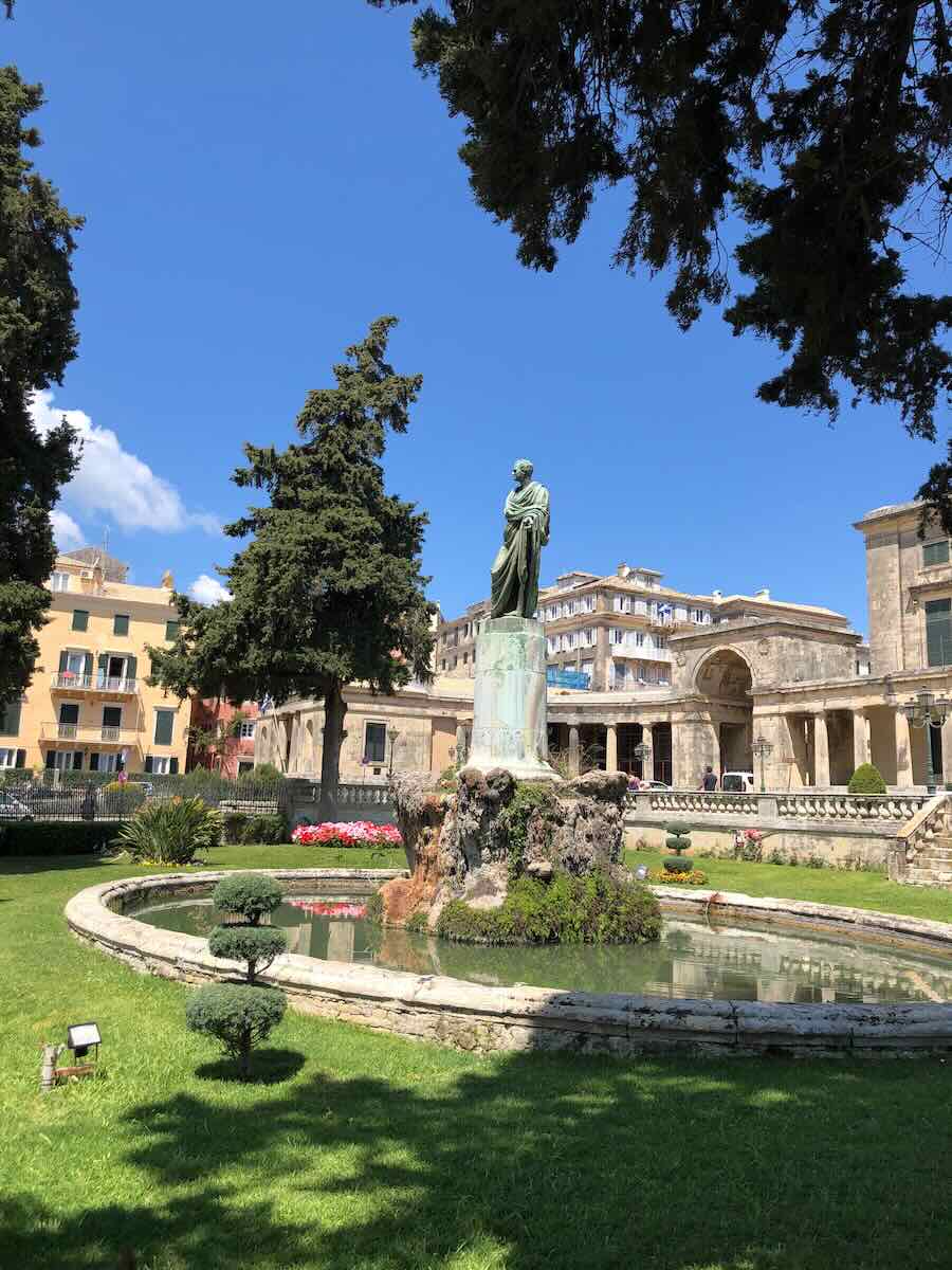 A scenic view of a statue in Corfu, Greece, surrounded by a circular fountain and lush green grass, with tall trees and historic buildings in the background under a clear blue sky.