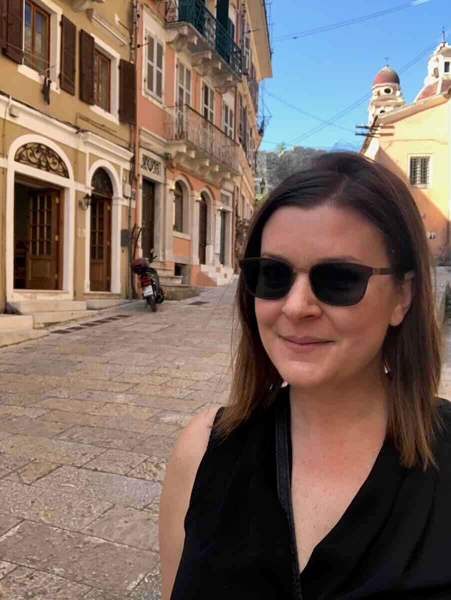 Melissa wearing sunglasses and a black top smiles while standing on a stone-paved street in Corfu, Greece. The background features pastel-colored buildings with balconies, a parked scooter, and a church dome visible in the distance.