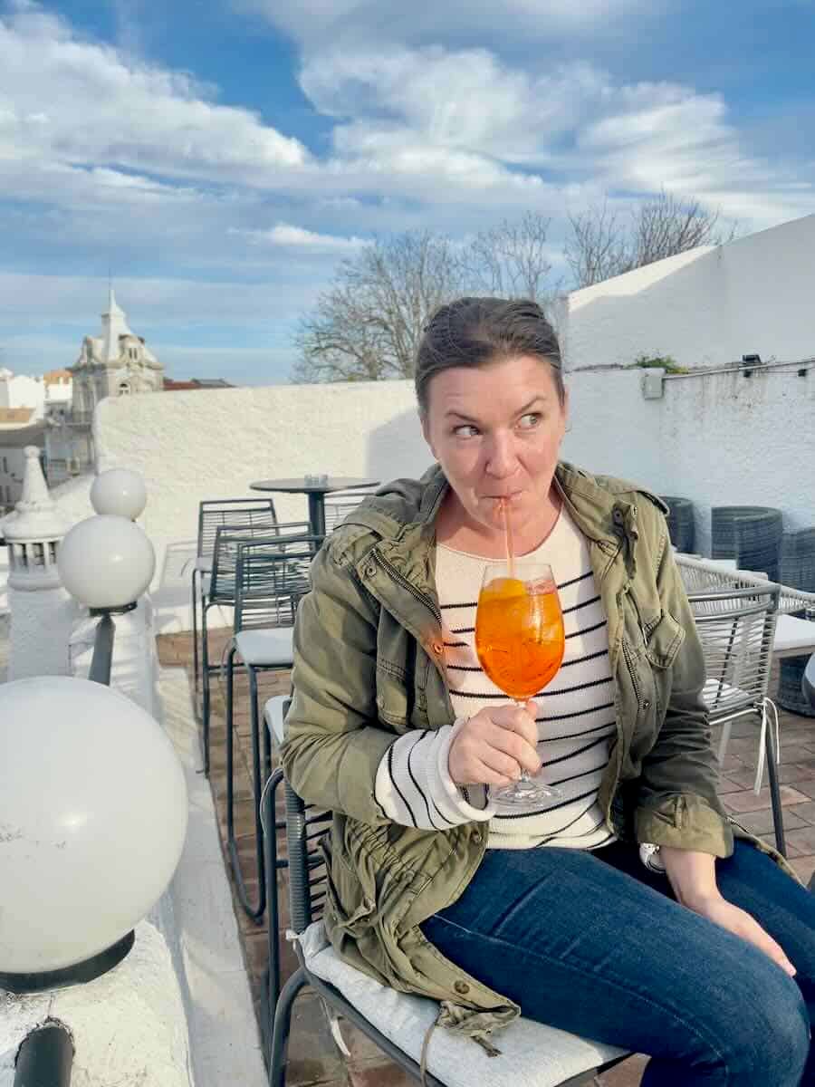 Melissa enjoying an Aperol spritz on a rooftop terrace in Faro, Portugal. The scene features whitewashed walls, modern outdoor furniture, and a partially cloudy blue sky with historic buildings visible in the background.