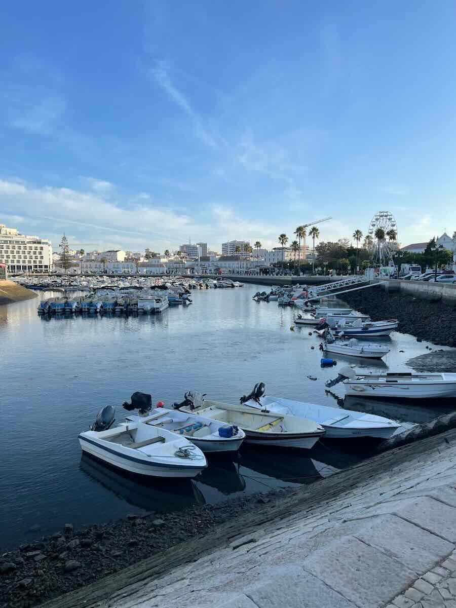 A serene harbor scene in Faro, Portugal, featuring small boats docked along the water, a backdrop of white buildings, palm trees, and a ferris wheel under a clear blue sky.