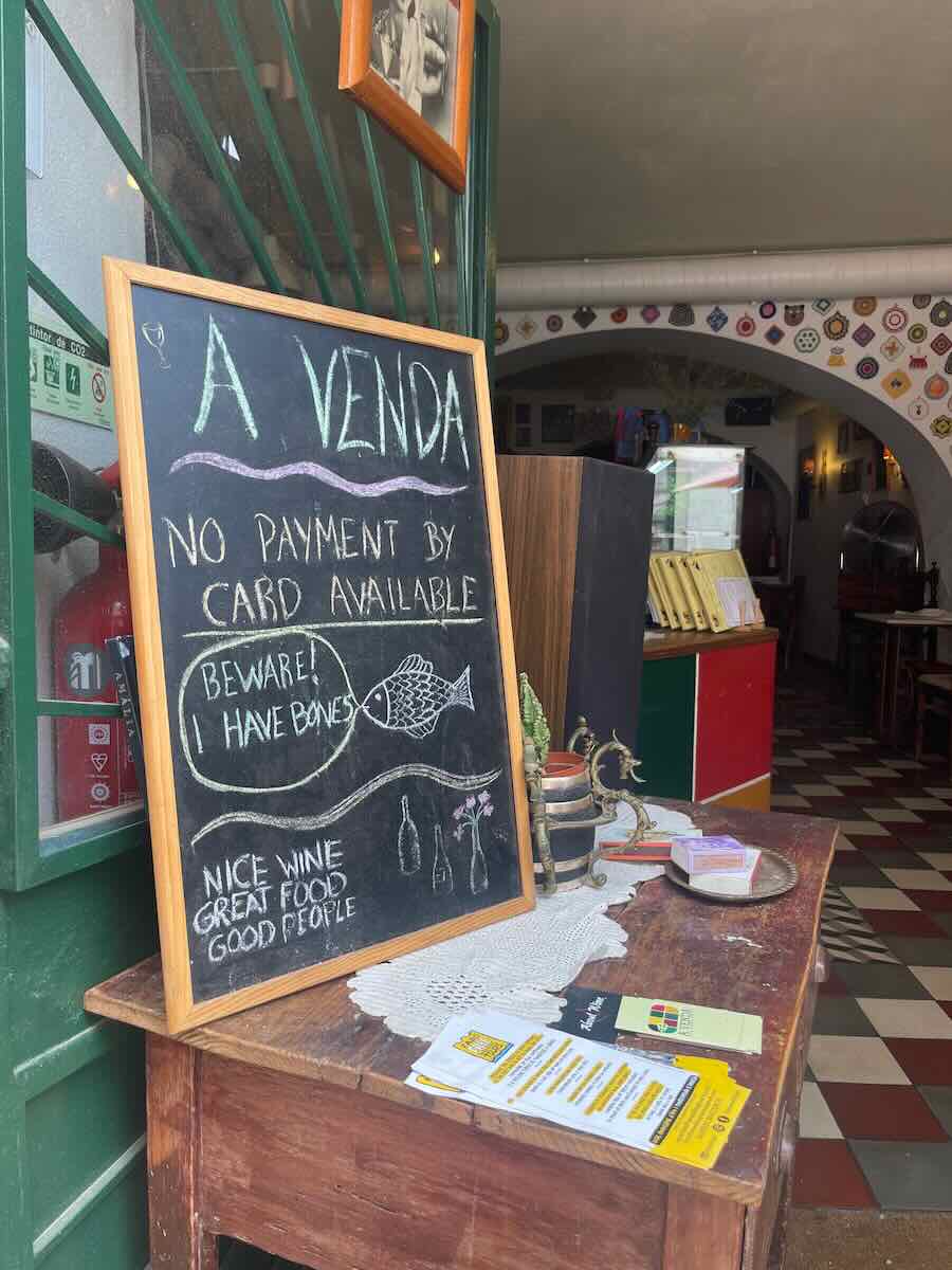 A chalkboard sign outside a restaurant called "A Venda" in Faro, Portugal, with a humorous message about the fish having bones and a reminder that they only accept cash payments.