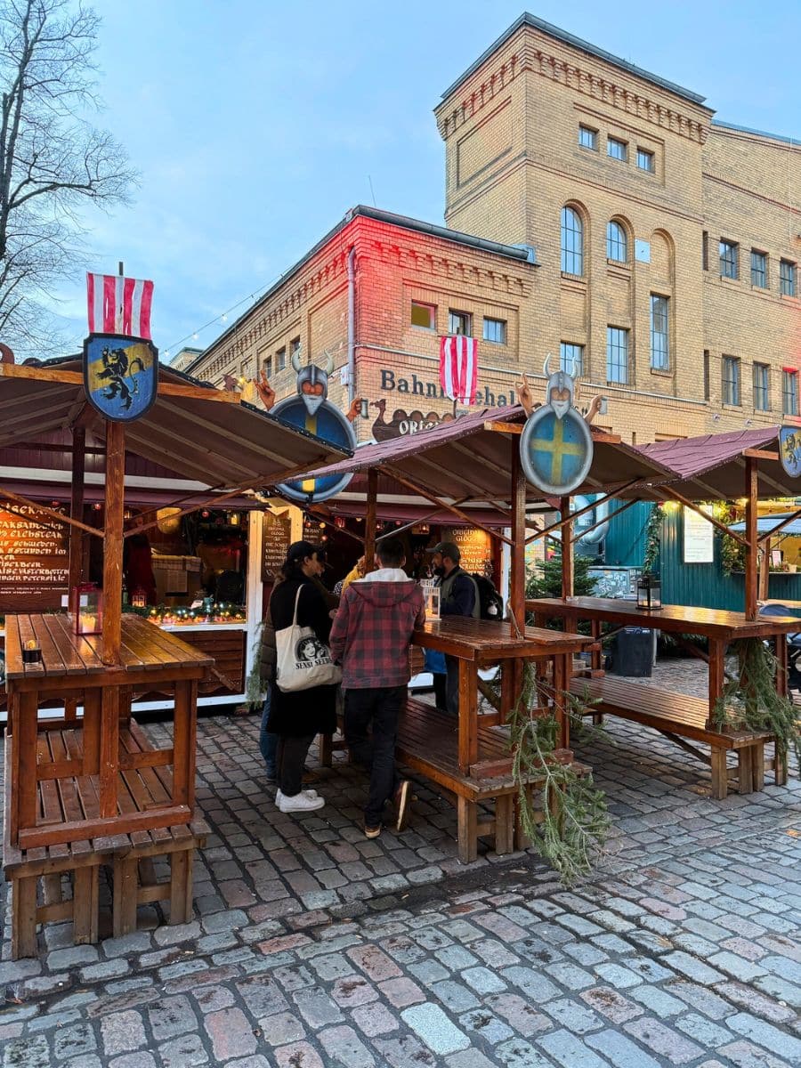 A group of people gathered around wooden tables under small covered stalls with Viking-themed decorations, set in a cobblestone courtyard at a Berlin Christmas market.