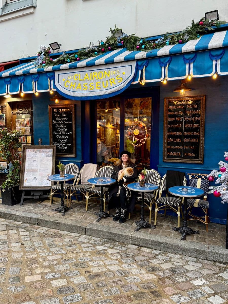 A Parisian café with blue awnings, outdoor seating, and festive decorations. Melissa in a black coat and beret holds Teddy, a fluffy dog while seated at one of the café's charming tables.