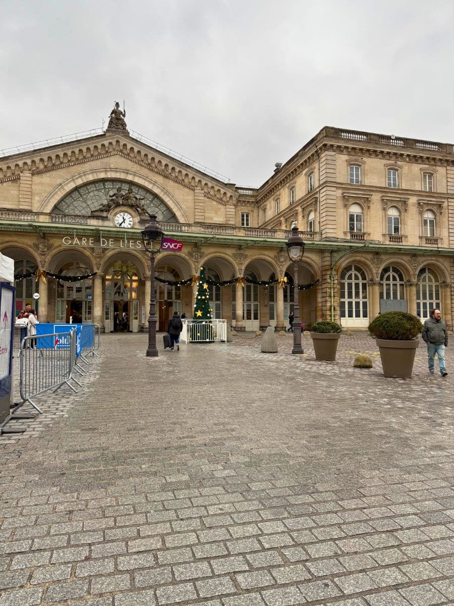 The exterior of Gare de l'Est in Paris, a historic train station with a large arched glass window and intricate architectural details. The cobblestone forecourt features potted plants and festive holiday decorations, including garlands and a Christmas tree. Passersby and travelers can be seen near the entrance.