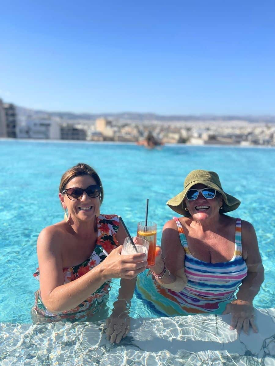 Two women enjoying drinks in a rooftop infinity pool with a cityscape and clear blue sky in the background, smiling and wearing colorful swimsuits and sunglasses.