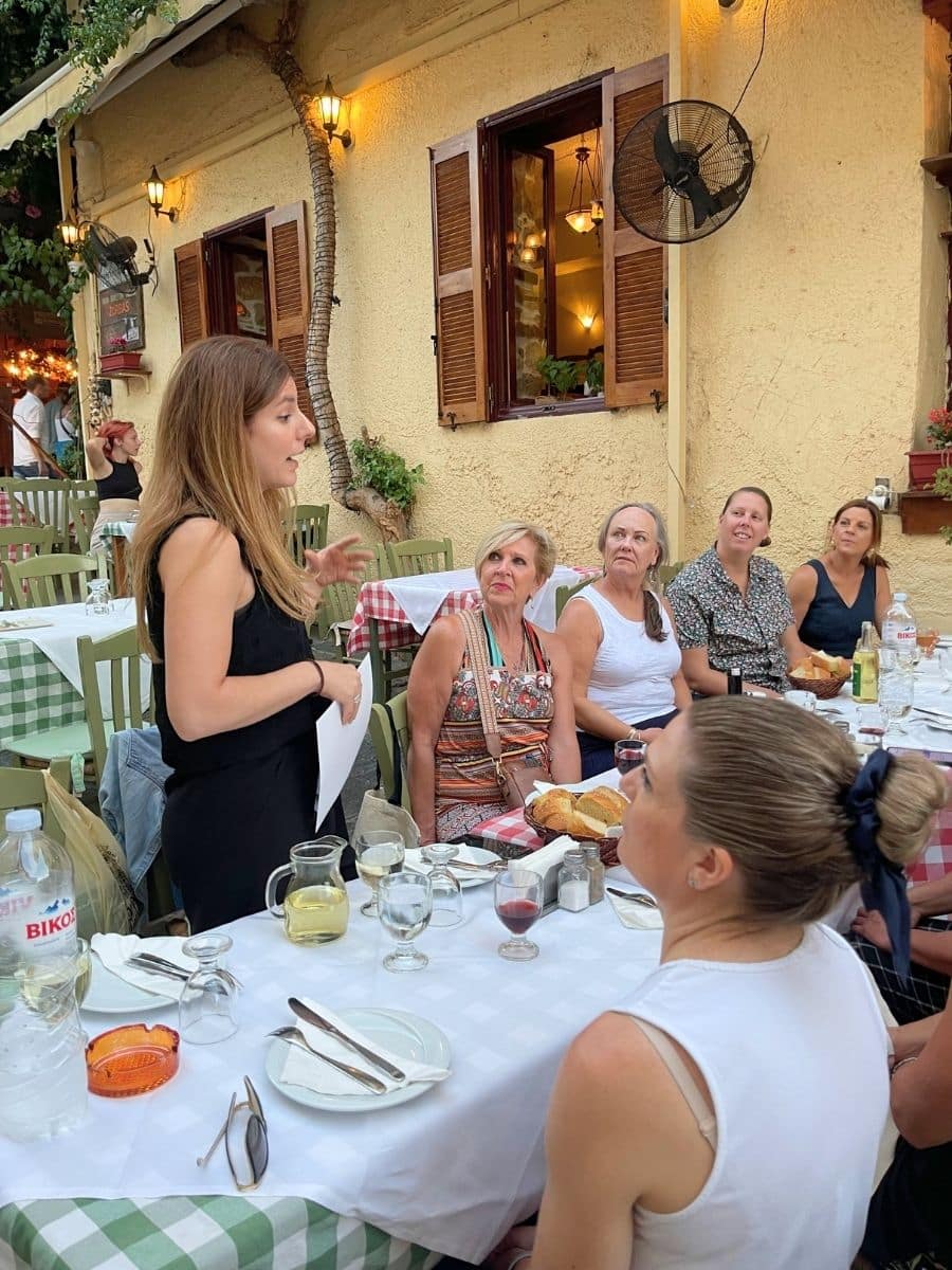 A woman standing and speaking to a group of women seated at an outdoor restaurant table, with plates, glasses, and a relaxed Mediterranean atmosphere in the background.
