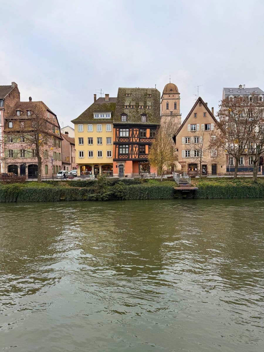 A serene riverside view in Strasbourg, showcasing charming half-timbered and traditional European houses with colorful facades. The reflective water adds a tranquil ambiance, while the historic architecture and cozy atmosphere capture the essence of Alsace's timeless beauty.