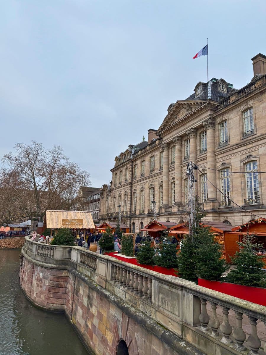 A closer view of a historic riverside building in Strasbourg, adorned with a French flag, featuring festive Christmas market stalls, evergreen trees, and a vibrant holiday atmosphere.
