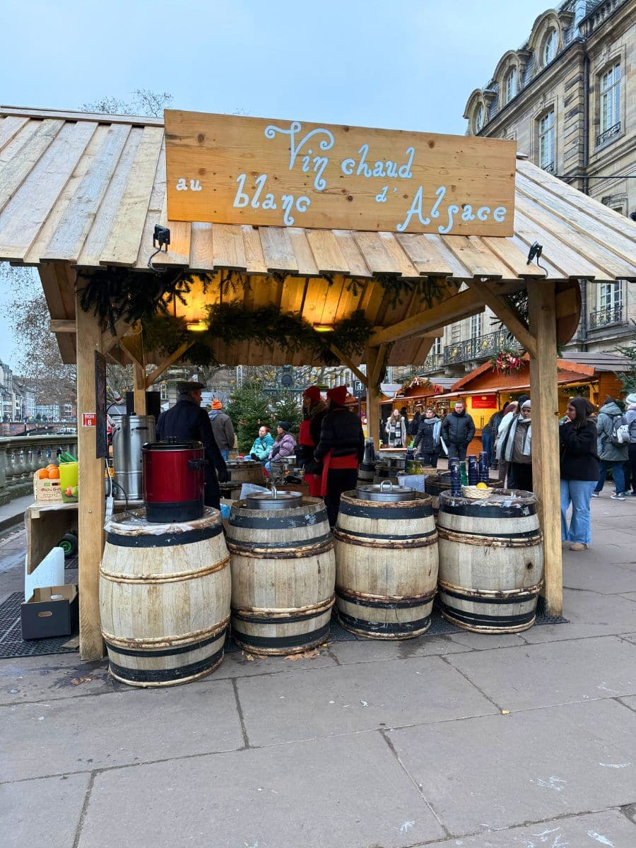 A charming wooden stall at the Strasbourg Christmas Market offers "Vin Chaud au Blanc d'Alsace," written in white cursive on a wooden sign above the stand. The stall is decorated with holiday greenery and features barrels as part of the display. Visitors in warm clothing gather around, with the backdrop of historic architecture and a festive atmosphere.