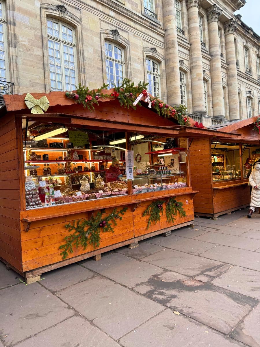 A charming wooden stall at the Strasbourg Christmas Market is adorned with festive garlands, ornaments, and candy cane decorations. It offers an array of sweet treats and handcrafted goods, with neatly displayed items visible through large glass windows. A historic stone building with grand columns and arched windows serves as a striking backdrop.