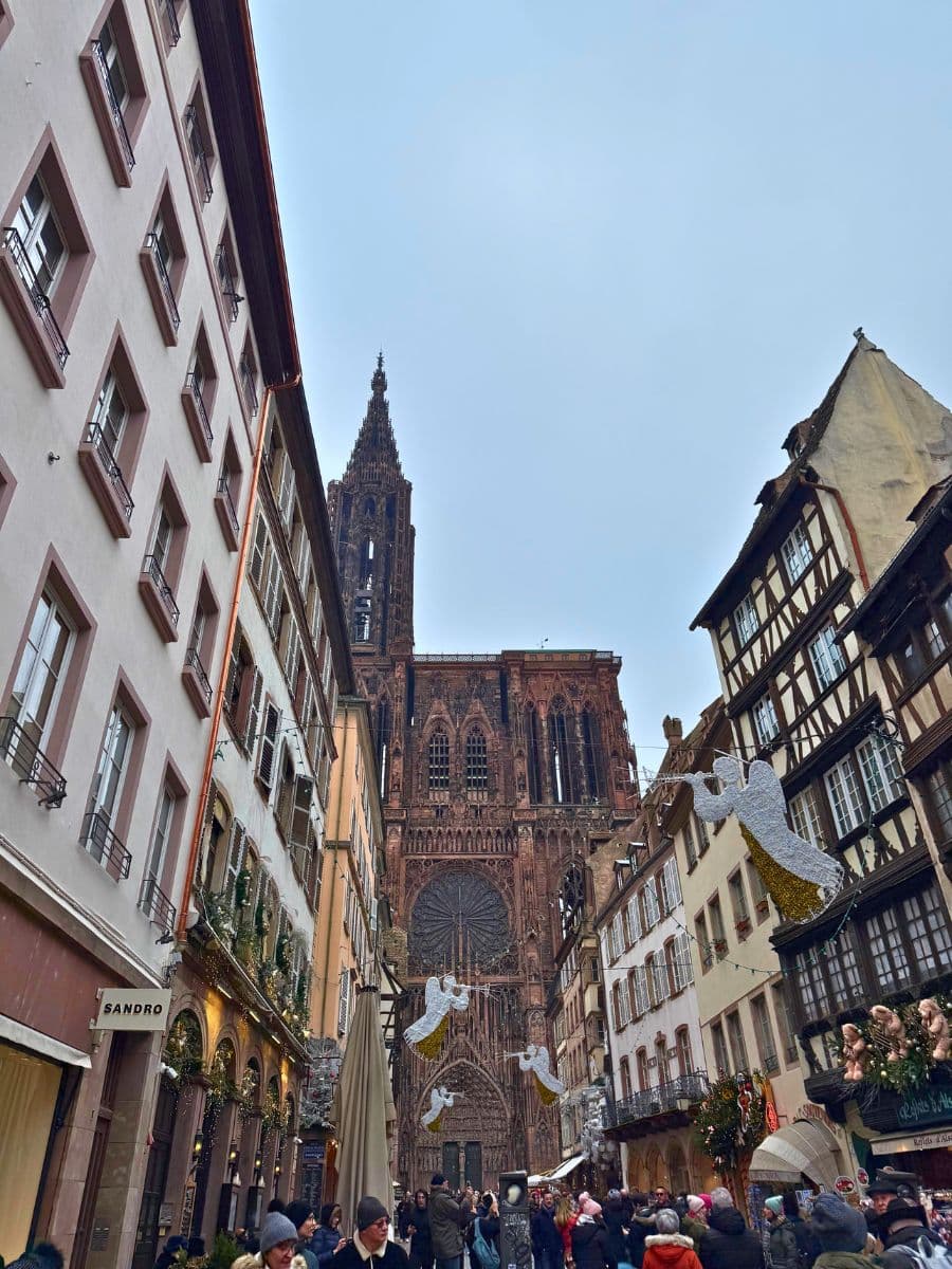 A bustling street in Strasbourg leading to the majestic Strasbourg Cathedral, framed by half-timbered and classic European-style buildings. The street is adorned with festive decorations, including angel-shaped ornaments with trumpets. The lively crowd adds to the holiday spirit, while the intricate architecture of the cathedral dominates the skyline under a pale winter sky.