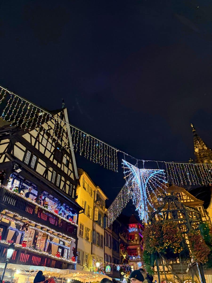 A festive scene at the Strasbourg Christmas Market featuring beautifully illuminated half-timbered buildings adorned with holiday decorations. Twinkling lights and intricate displays hang across the street, creating a magical atmosphere under the dark evening sky. Visitors are seen enjoying the lively surroundings.