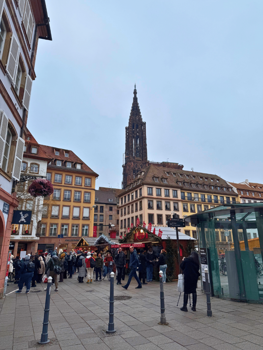 This image captures a lively Christmas market in Strasbourg with a festive atmosphere. The iconic spire of Strasbourg Cathedral stands tall in the background, creating a stunning contrast against the historic half-timbered architecture surrounding the square. Crowds gather around charming wooden market stalls adorned with decorations, showcasing a variety of holiday treats and crafts. The hanging flower baskets and the blend of modern and historic details add to the charm of this bustling holiday scene.
