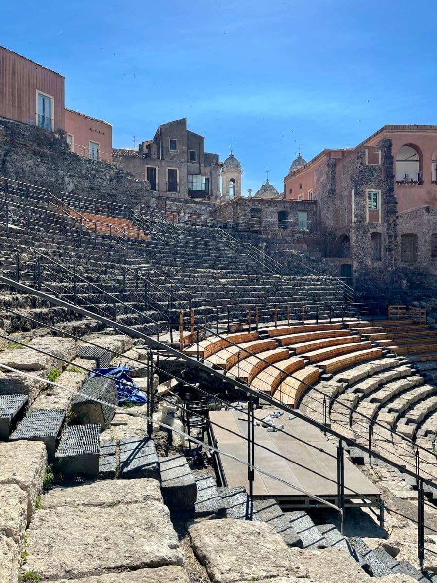 A closer view of the ancient Roman amphitheater in Catania, Sicily, showcasing stone seating, metal railings, and a modern stage setup. The ruins are surrounded by historic buildings with church domes in the background under a clear blue sky.