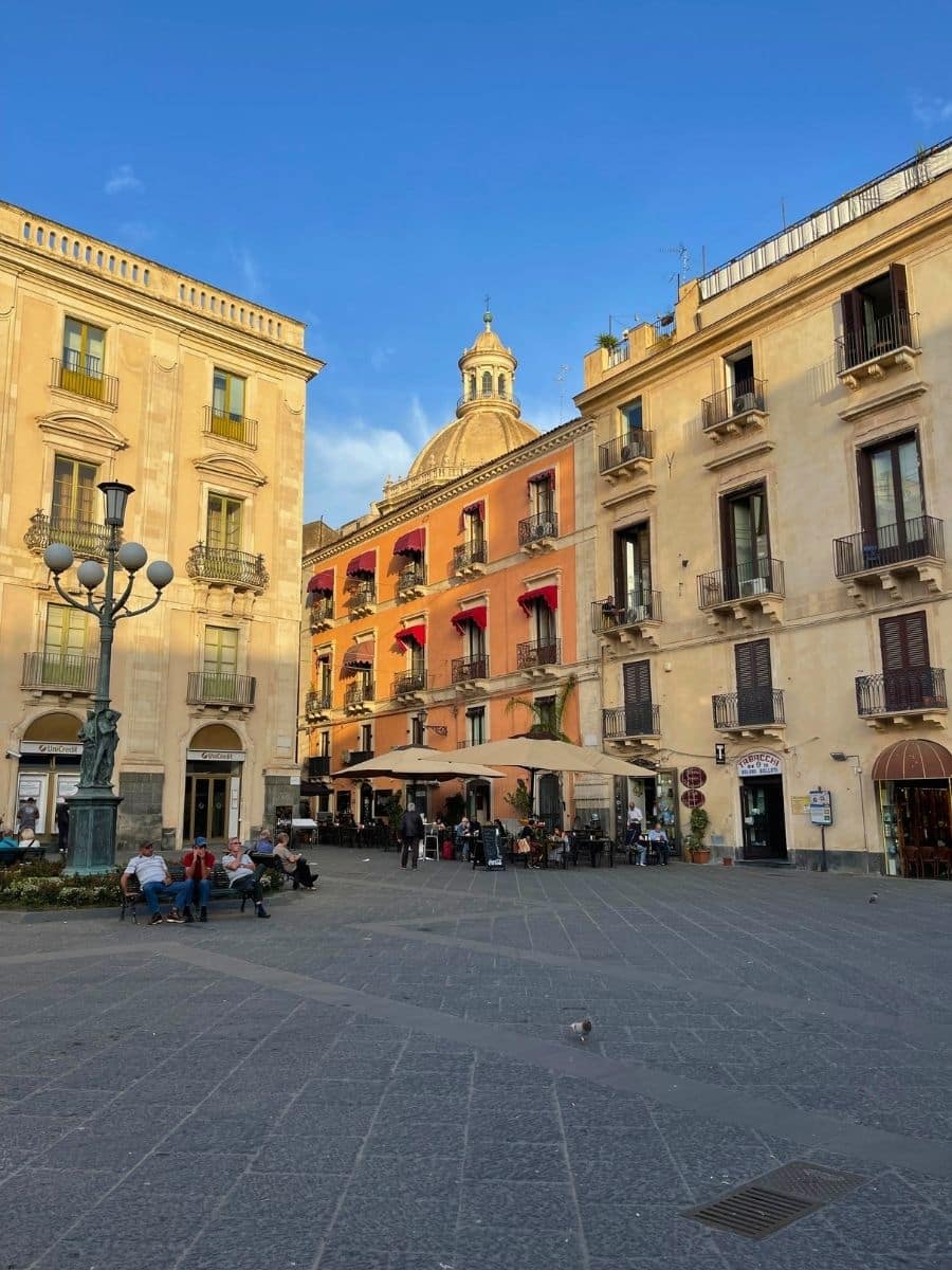 Piazza Università in Catania, Sicily, with its light-colored stone buildings, decorative street lamps, and a central area where people are sitting and relaxing. The prominent dome of the university building is visible in the background.