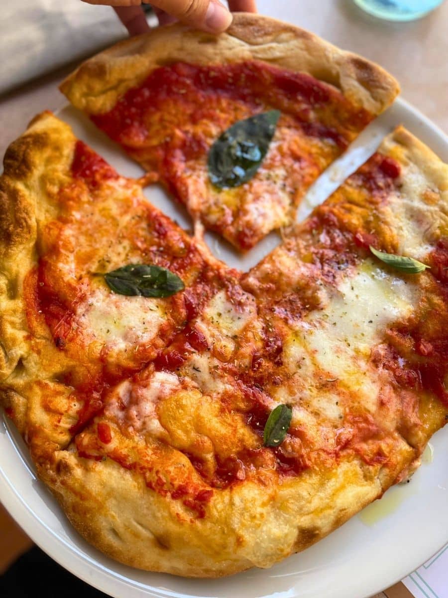 A close-up of a freshly baked Margherita pizza with a golden, crispy crust, topped with melted mozzarella, tomato sauce, and fresh basil leaves. A hand is lifting a slice, revealing the gooey cheese and flavorful toppings on a white plate.
