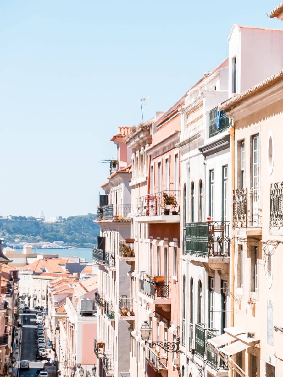A sunlit street in Lisbon, Portugal, lined with colorful pastel buildings featuring traditional iron balconies. The street slopes downward towards the Tagus River, with a scenic view of the water and distant green hills in the background.